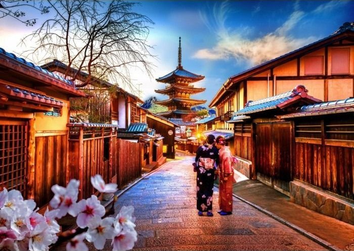 Two women in kimonos stand on a traditional Japanese stone street with a pagoda in the distance.