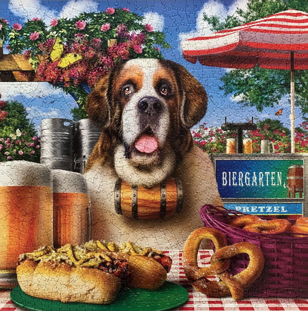 A Saint Bernard dog wearing a small barrel collar sits at a sunny outdoor beer garden table with beer and pretzels.
