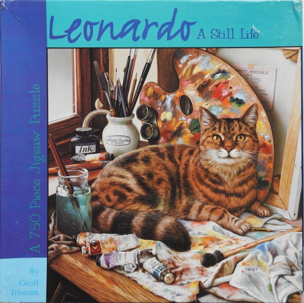 A brown tabby cat lounging on an artist's workspace cluttered with paint tubes, brushes, and a palette.