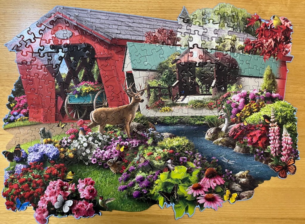 A whimsical scene of a red covered bridge over a stream in a flower-filled garden with a deer.