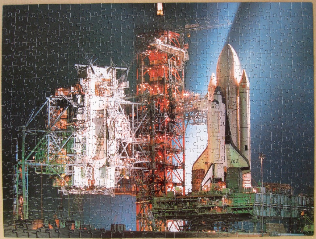 A space shuttle perched on a launch pad at night, illuminated by bright floodlights against a dark sky.