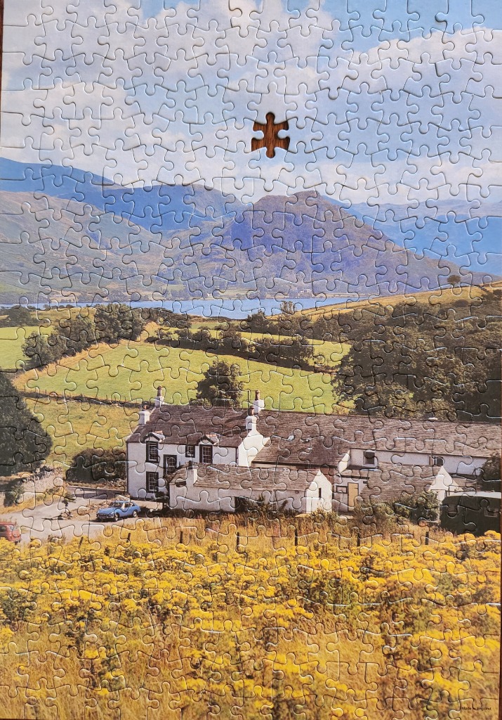 A landscape view of a white farmhouse in front of a blue lake and rolling mountains under a cloudy sky.