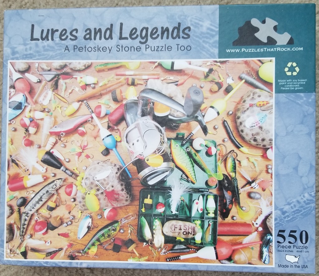 A cluttered collection of colorful fishing gear, lures, bobbers, and a reel on a wooden background with Petoskey stones.