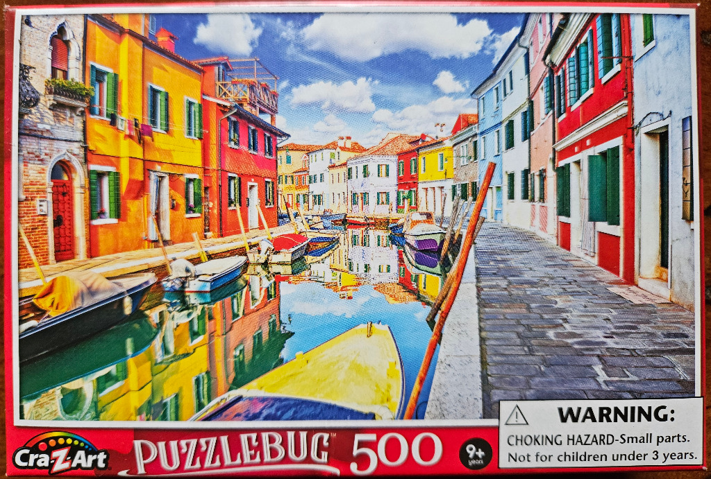 A sunny scene featuring a narrow canal lined with colorful, brightly painted buildings on the island of Burano, Venice.