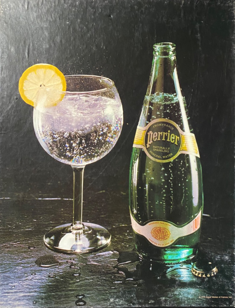 A green glass bottle of sparkling water next to a round goblet filled with the same, garnished with a lemon slice.