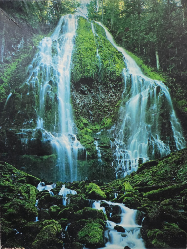A lush, tiered waterfall cascading down a moss-covered cliff face into a rocky stream.