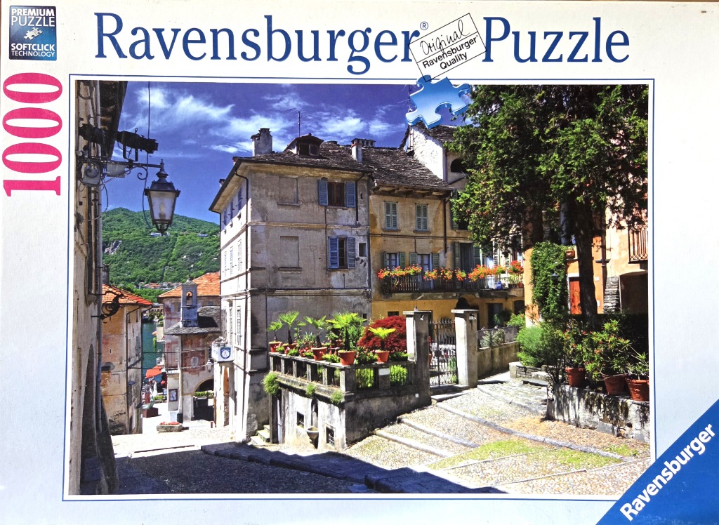 A sunny Italianate street scene with old stone buildings, potted plants, and a view towards a distant lake and mountains.