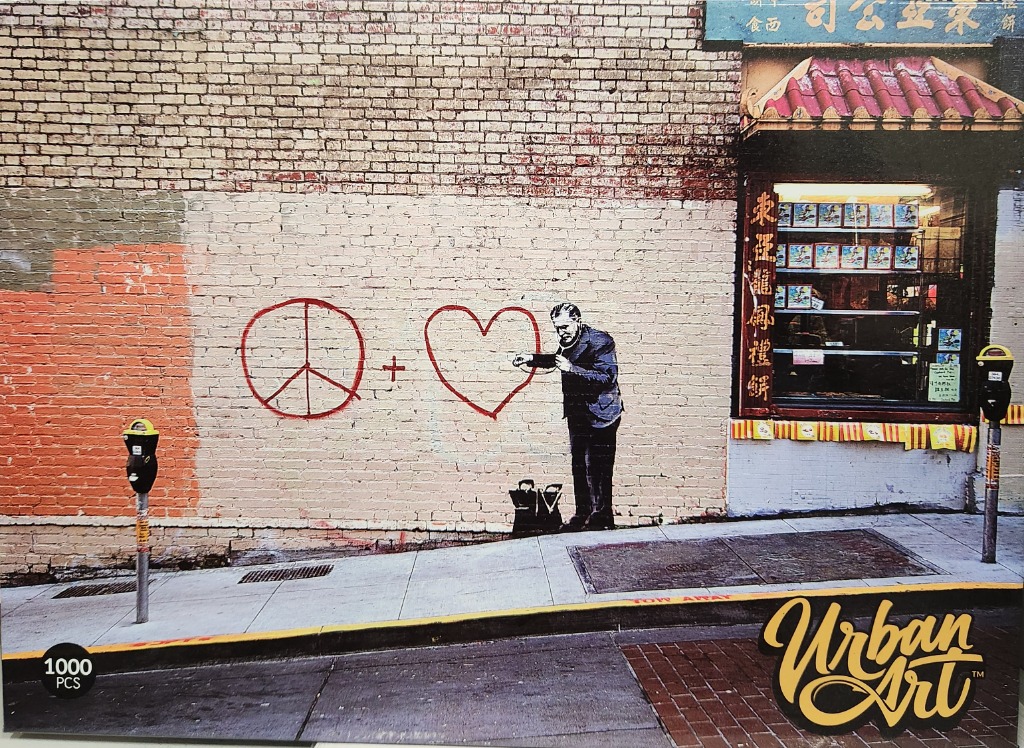 Street art mural of a man painting a peace sign and heart on a brick wall next to a storefront.