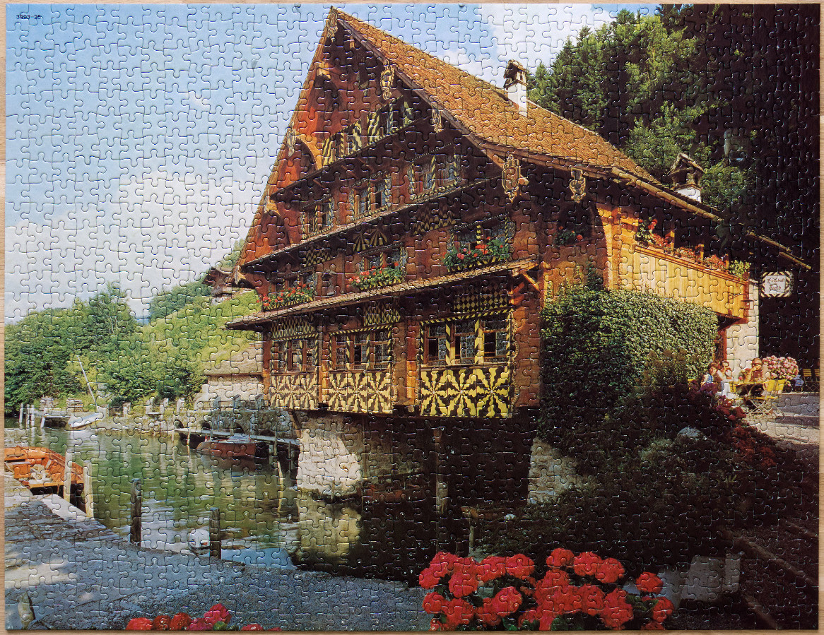 A traditional multi-story Swiss wooden house situated on the edge of a canal with vibrant flower boxes and greenery.