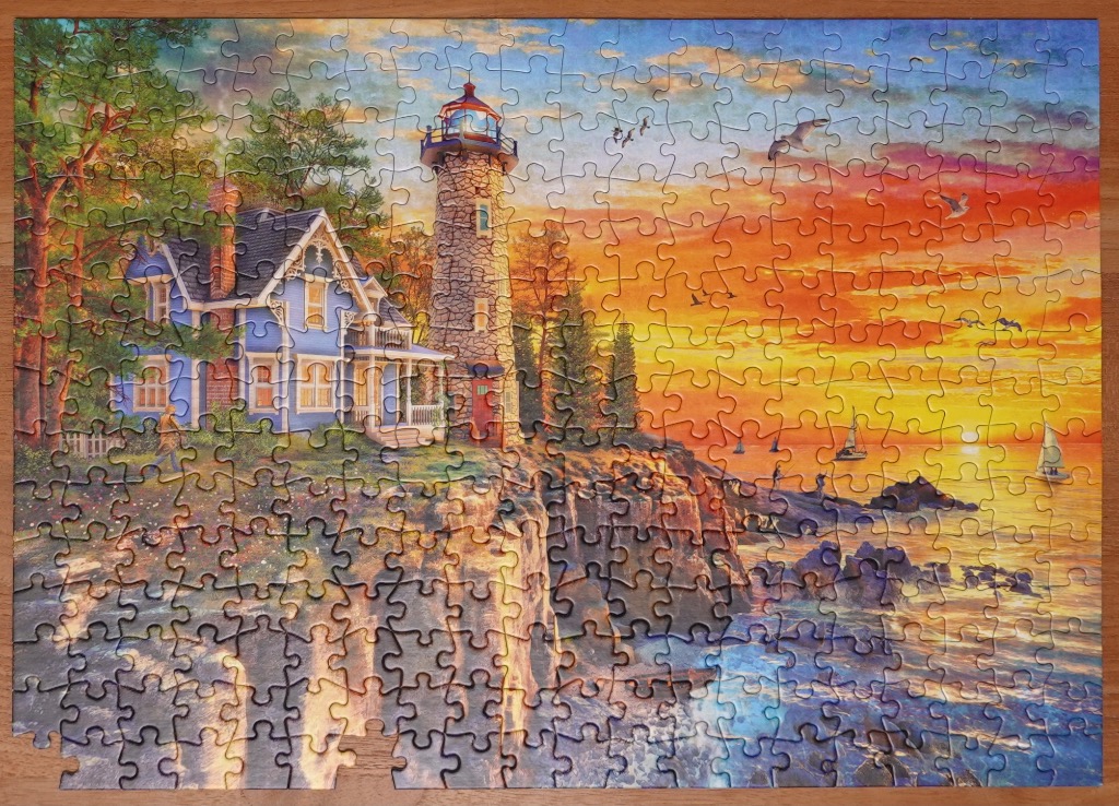 A lighthouse and victorian house on a cliff at sunset with sailboats on the water.