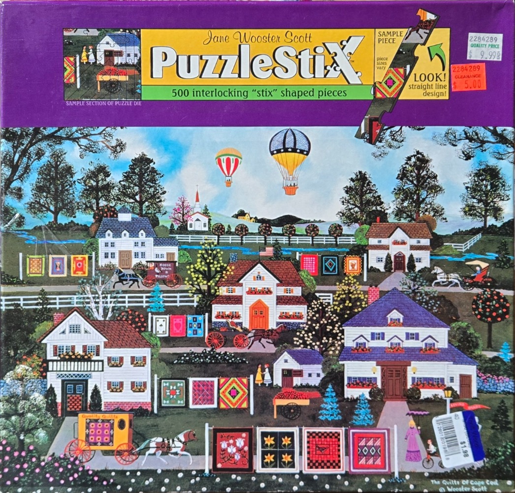 Folk art scene of houses in a rural landscape with quilts hung on fences and hot air balloons in the sky.