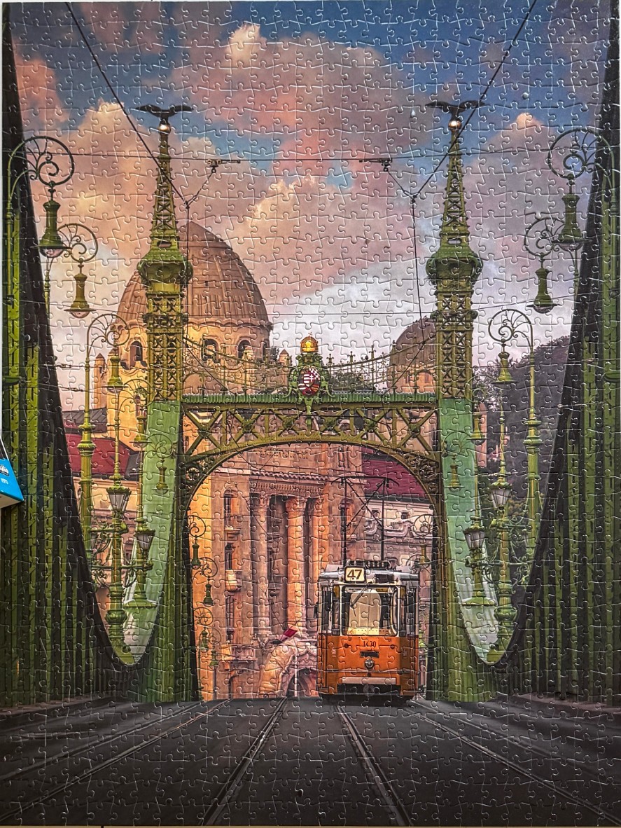 A view through the ornate green metal trusses of the Liberty Bridge in Budapest featuring a vintage orange tram.