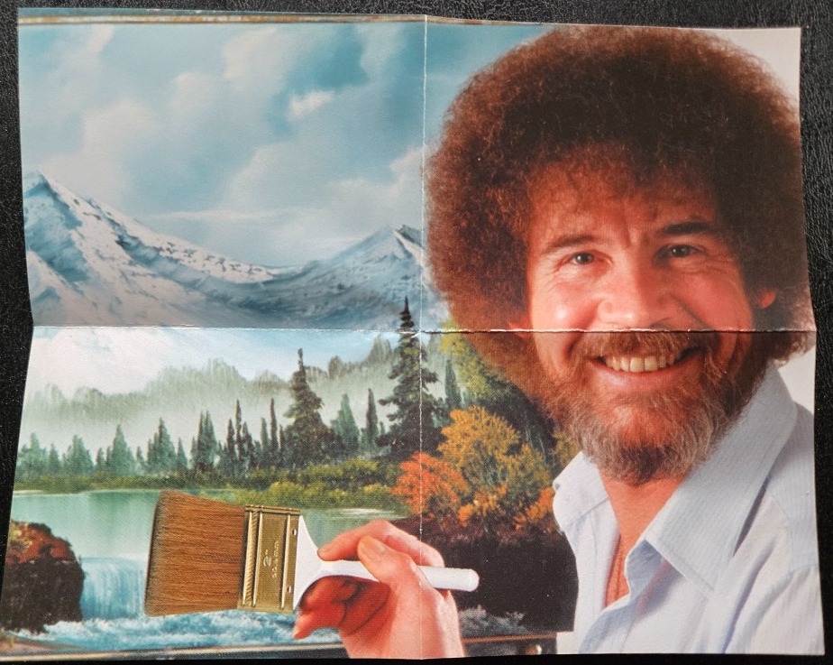 A smiling man with an afro and beard holding a paintbrush in front of a painted landscape.