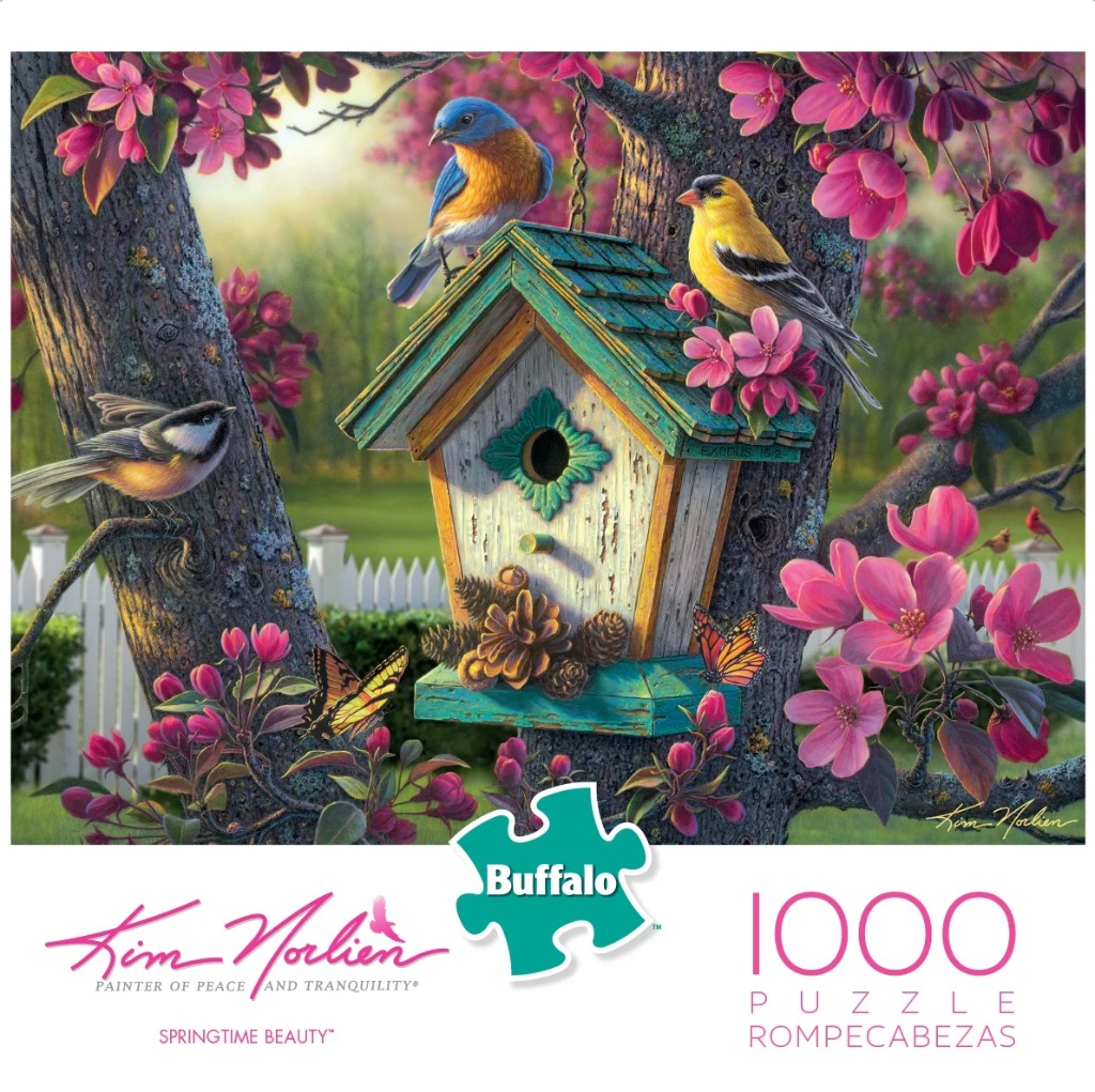 A bluebird and a goldfinch perched on a weathered wooden birdhouse surrounded by pink spring blossoms.