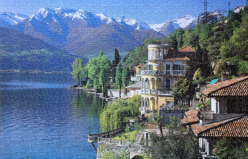 An elegant Italian villa on the shore of a deep blue lake with snow-capped mountains in the background.