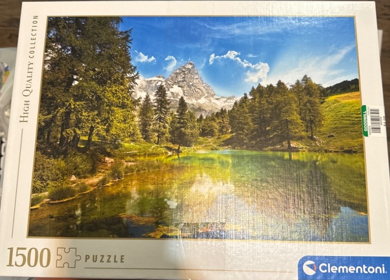 A clear mountain lake reflecting a jagged, snow-dusted peak surrounded by green trees and meadows.