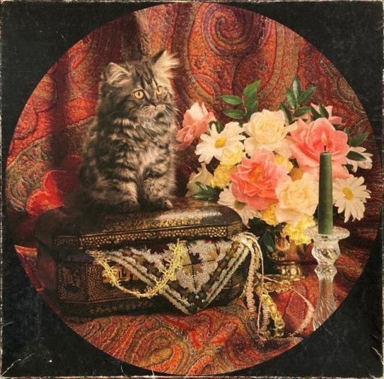 A fluffy brown tabby kitten sits on an ornate box next to a bouquet of flowers and a green candle.