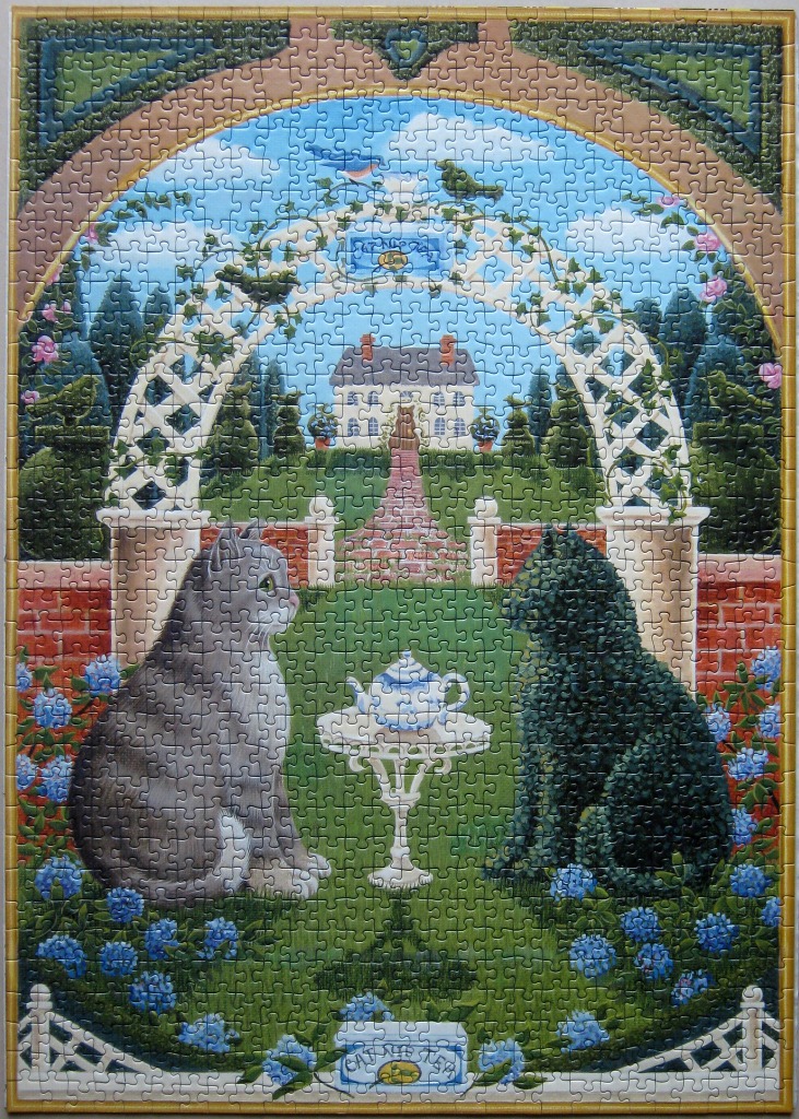 A grey cat and a black cat sit on a lawn facing a small table in a decorative garden.