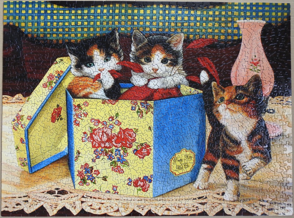 Three calico kittens in and around a floral decorative gift box on a lace tablecloth.