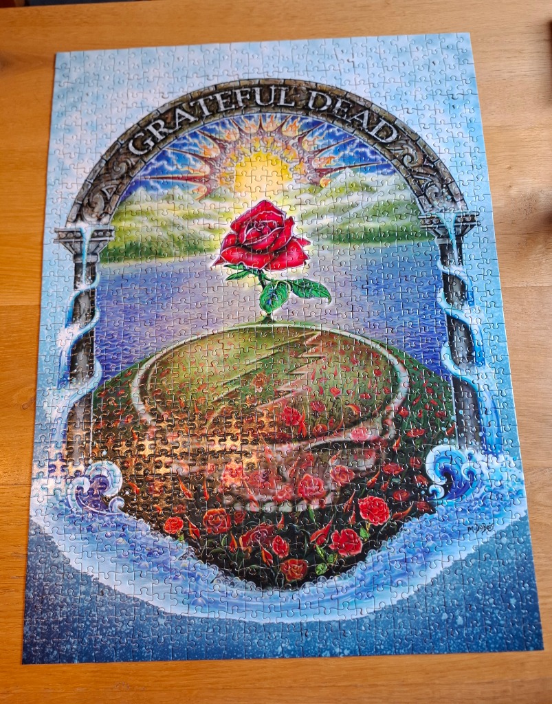 Surreal illustration of a single red rose rising from a grassy mound framed by a stone archway with the Grateful Dead logo.