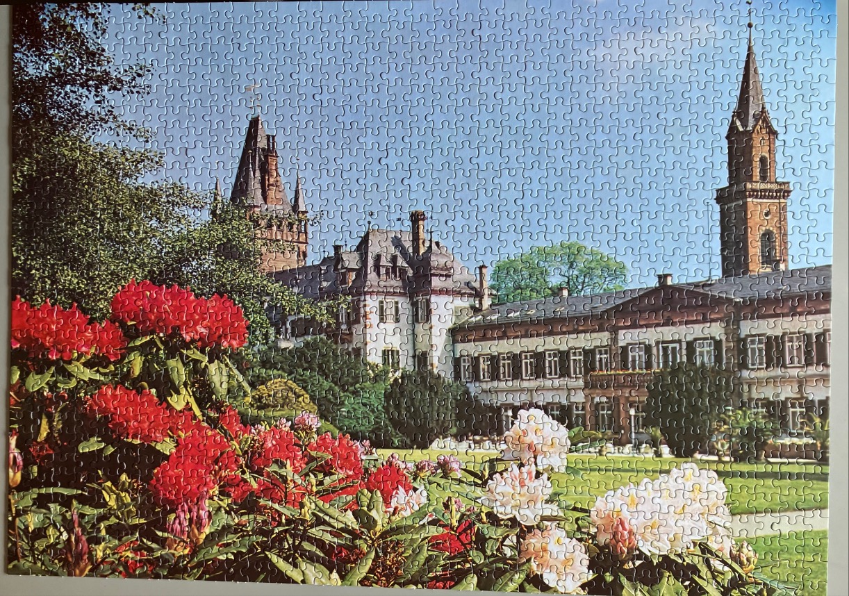 A sunny European castle with a prominent tower, surrounded by lush gardens featuring vibrant red and white rhododendrons.