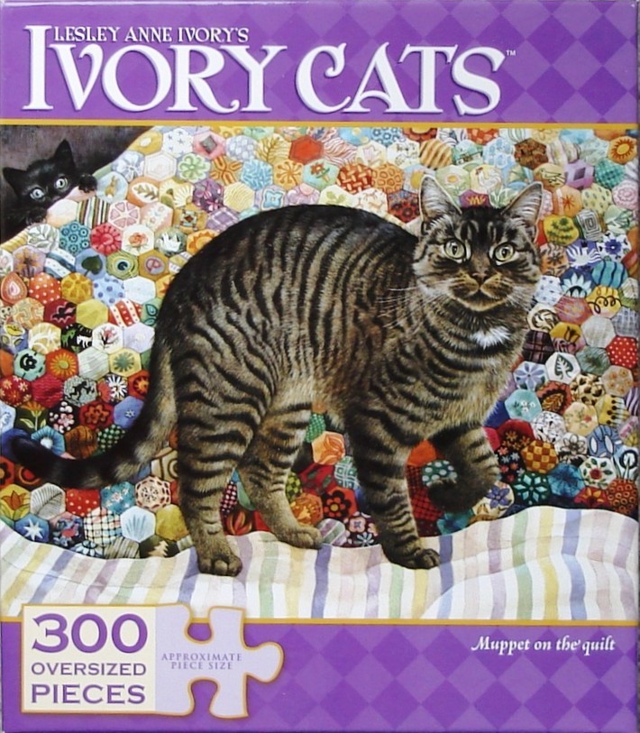 A tabby cat standing on a colorful hexagon-patterned quilt, with a small black kitten peeking from behind in the corner.