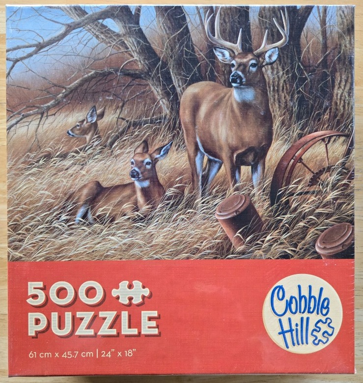 A buck and two does in a tall grass field near a rusted antique wagon wheel.