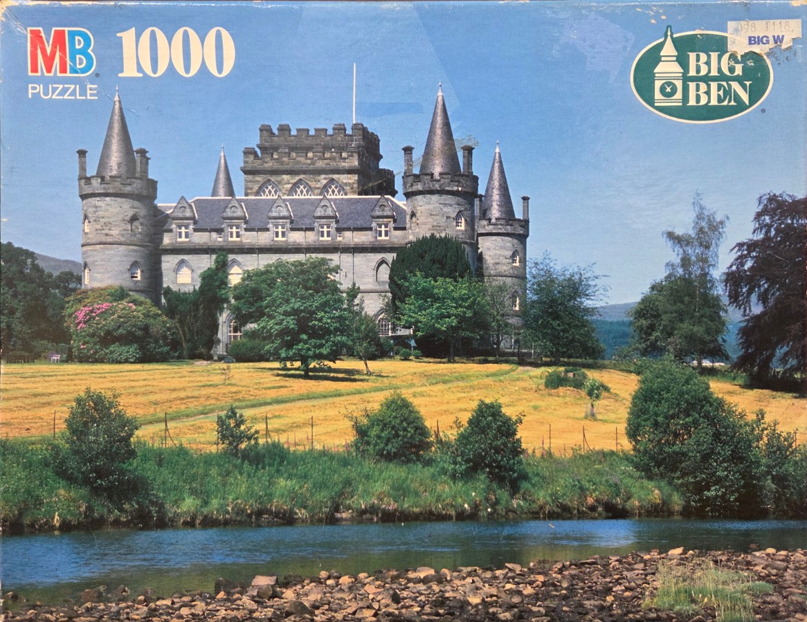 A photograph of Inveraray Castle in Scotland, featuring grey stone towers, a golden meadow, and a foreground river.
