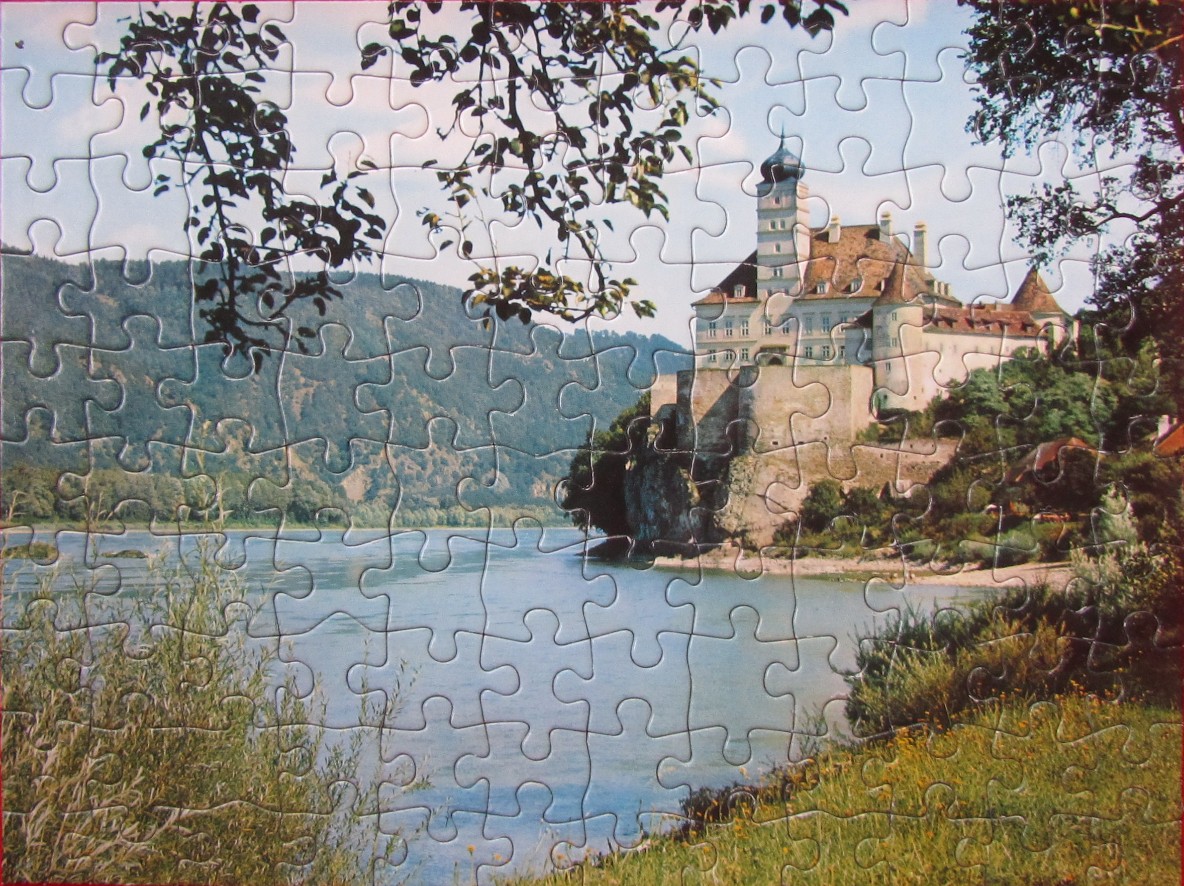 A European castle perched on a rocky cliff overlooking a wide river with forested hills in the background.