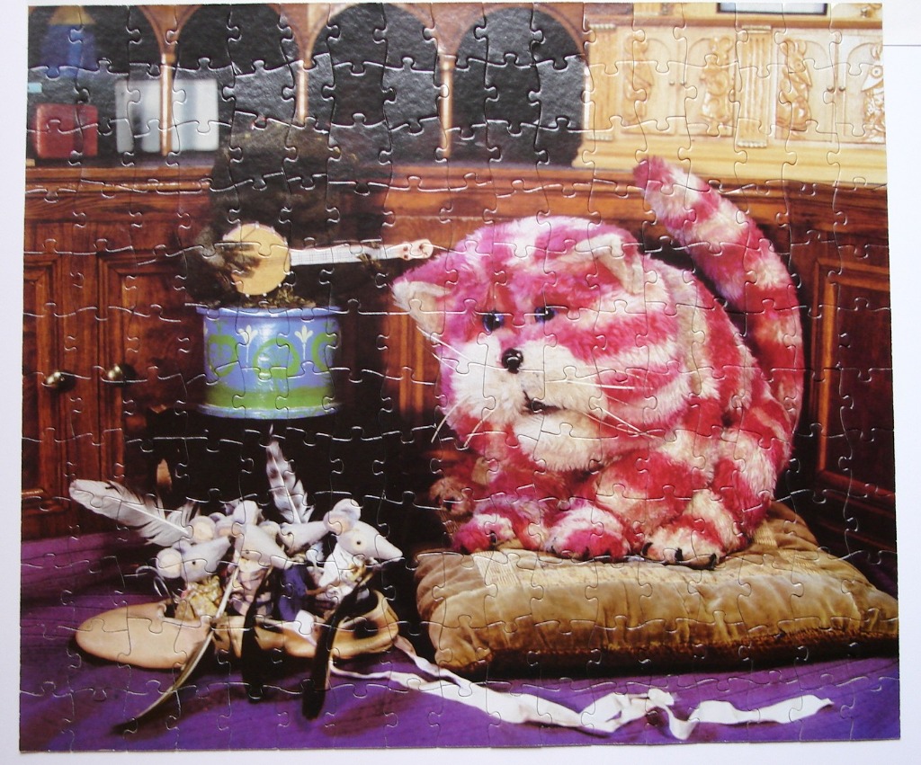 A whimsical plush pink-and-white striped cat sitting on a cushion in a room with dark wood furniture and toy mice.