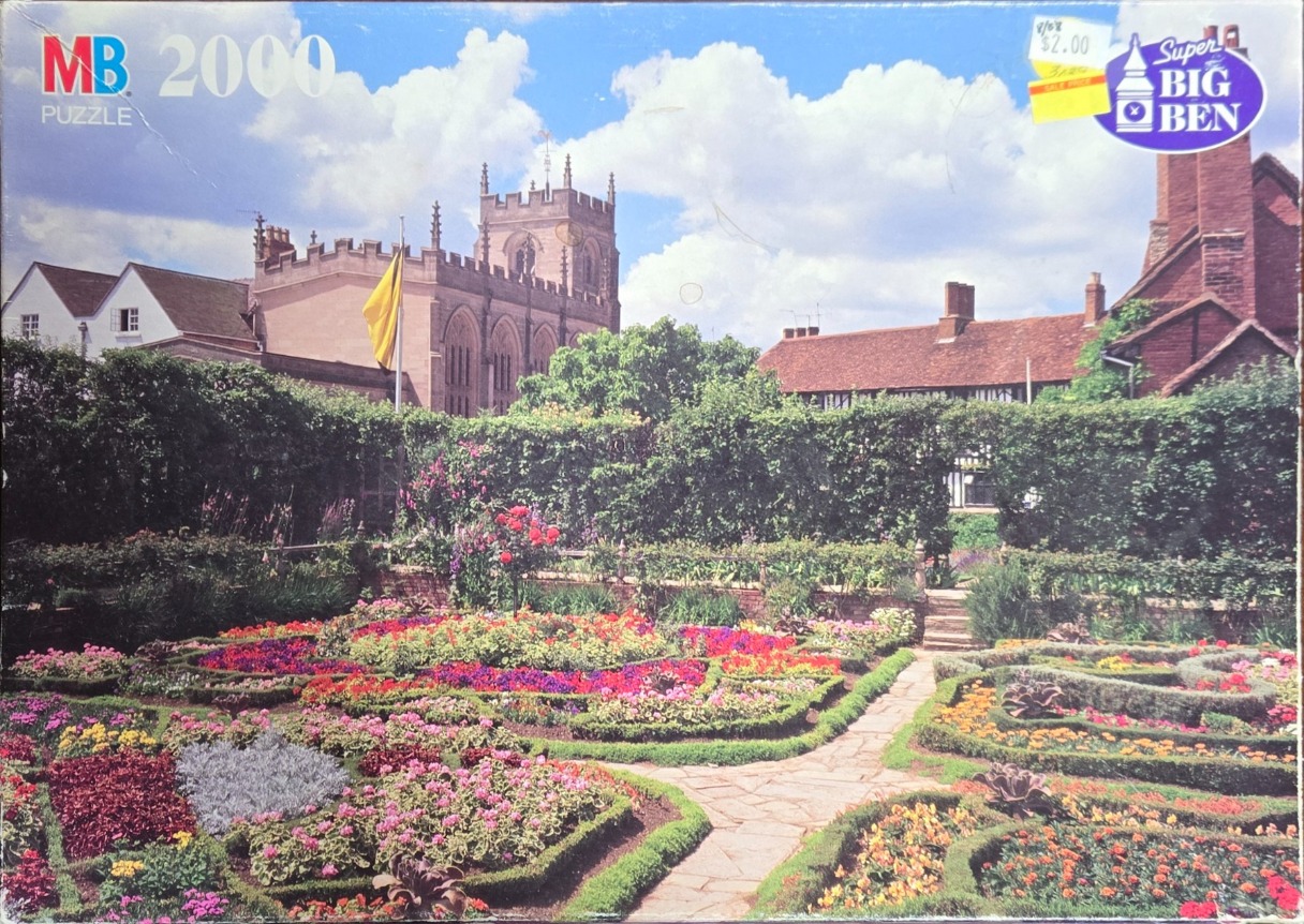 A lush, formal English garden with colorful flower beds in the foreground and a historic stone church in the background.