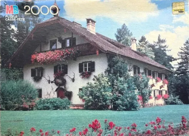 A traditional Alpine-style white farmhouse with red geraniums and a green lawn under a blue sky.
