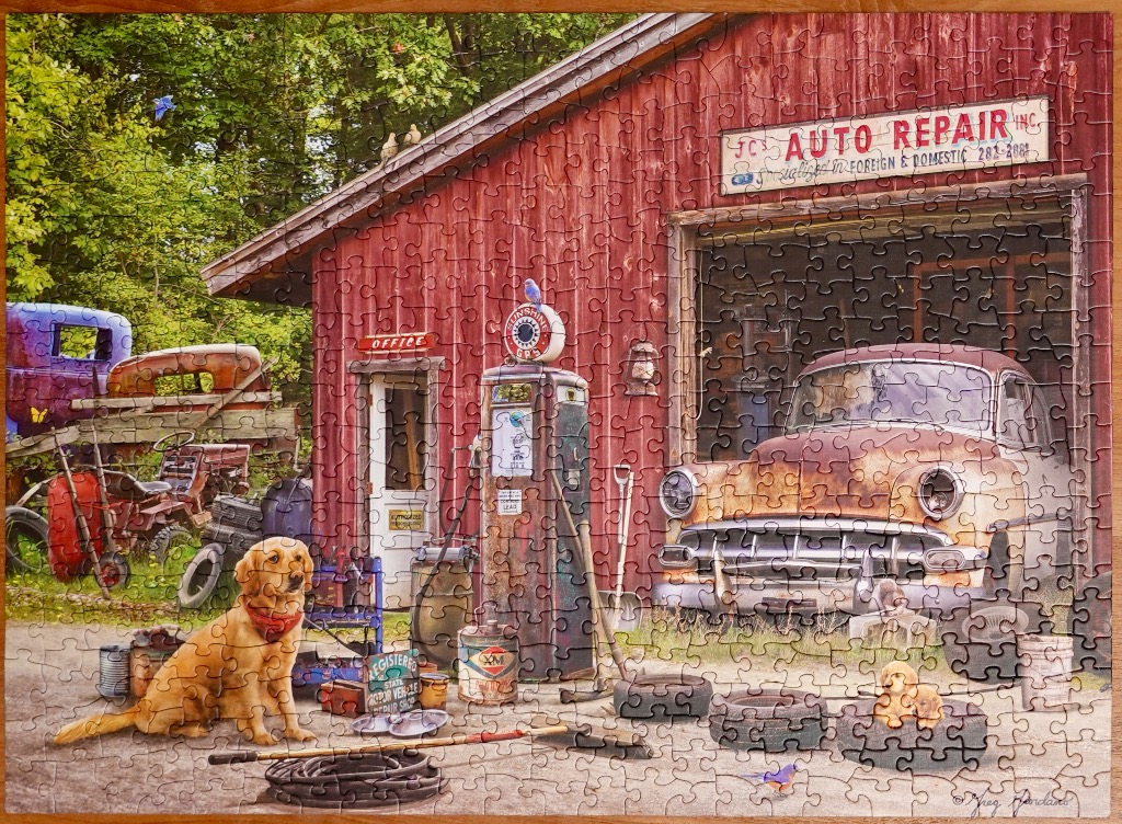 A golden retriever sits in front of a rustic red barn auto repair shop with a vintage car inside.