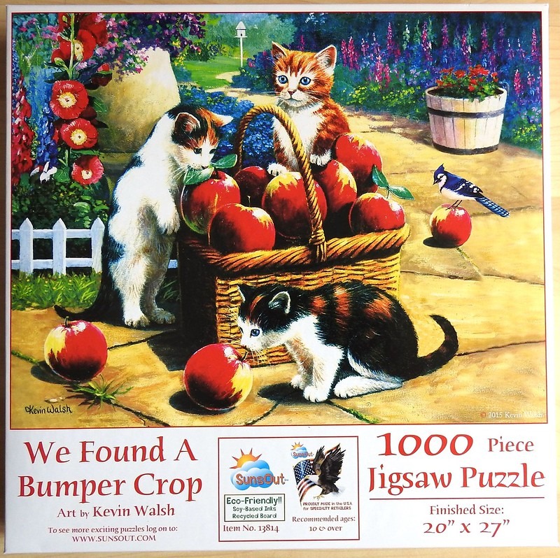 Three kittens playing with a wicker basket full of red apples in a sunlit garden.