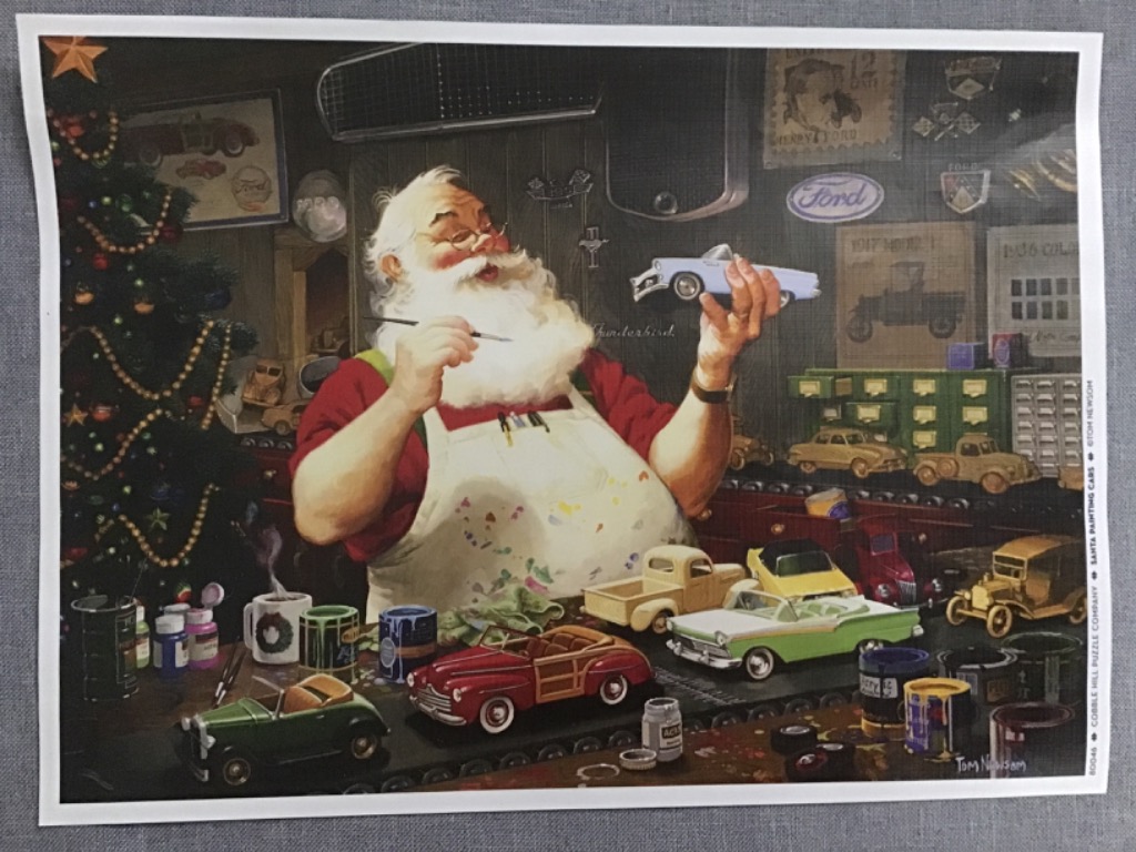 Santa Claus painting miniature toy cars at a workbench near a decorated Christmas tree.