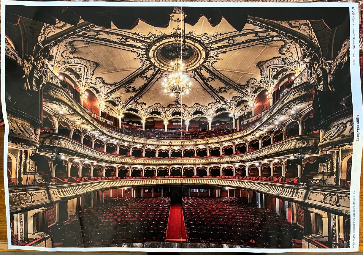 Wide-angle interior view of an ornate, historic multi-tiered opera house with red seating and a central chandelier.