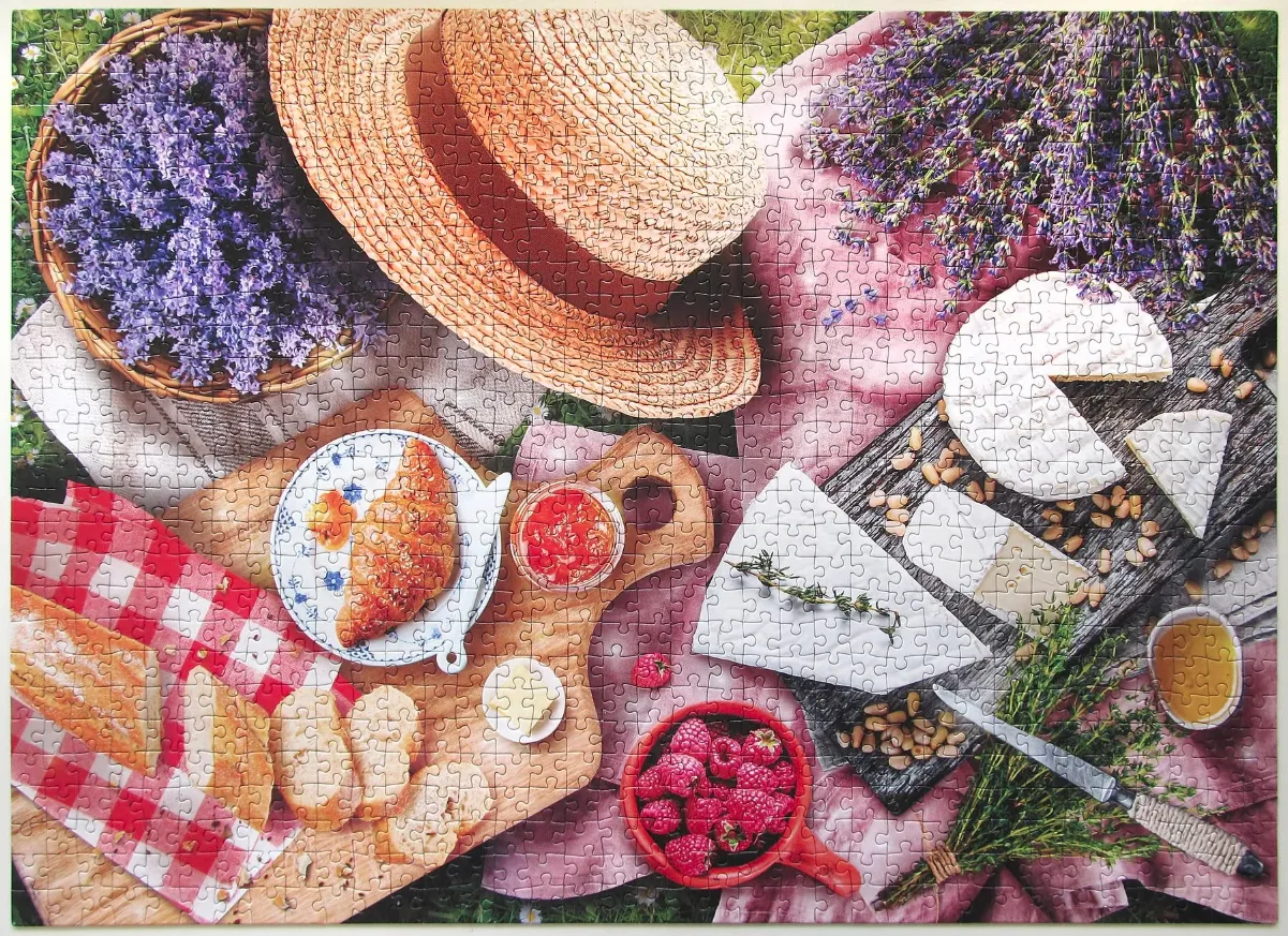 A top-down view of a rustic outdoor picnic featuring a straw hat, lavender, cheeses, bread, and raspberries.