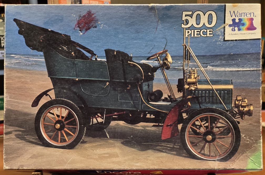 A vintage dark green touring car parked on a sandy beach by the ocean.