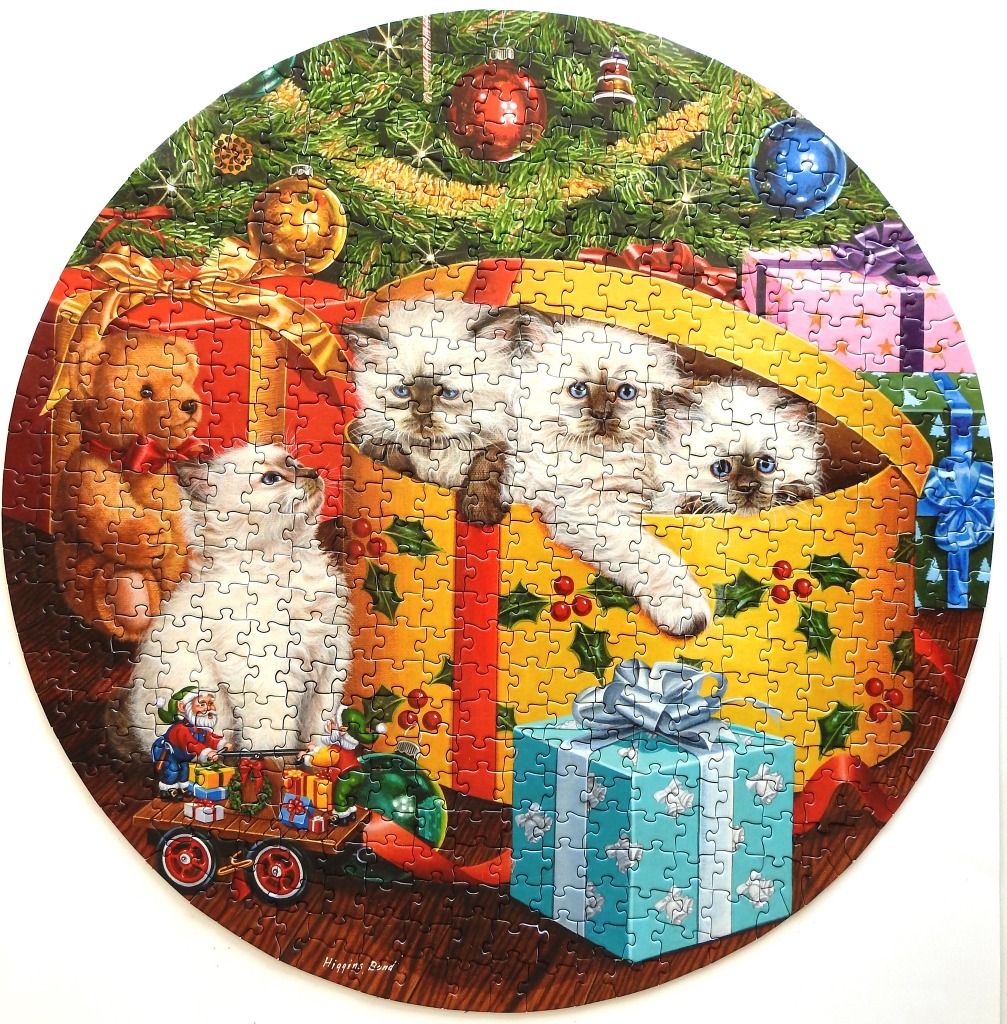 Three fluffy kittens peeking out of a large yellow Christmas gift box under a tree surrounded by toys and presents.