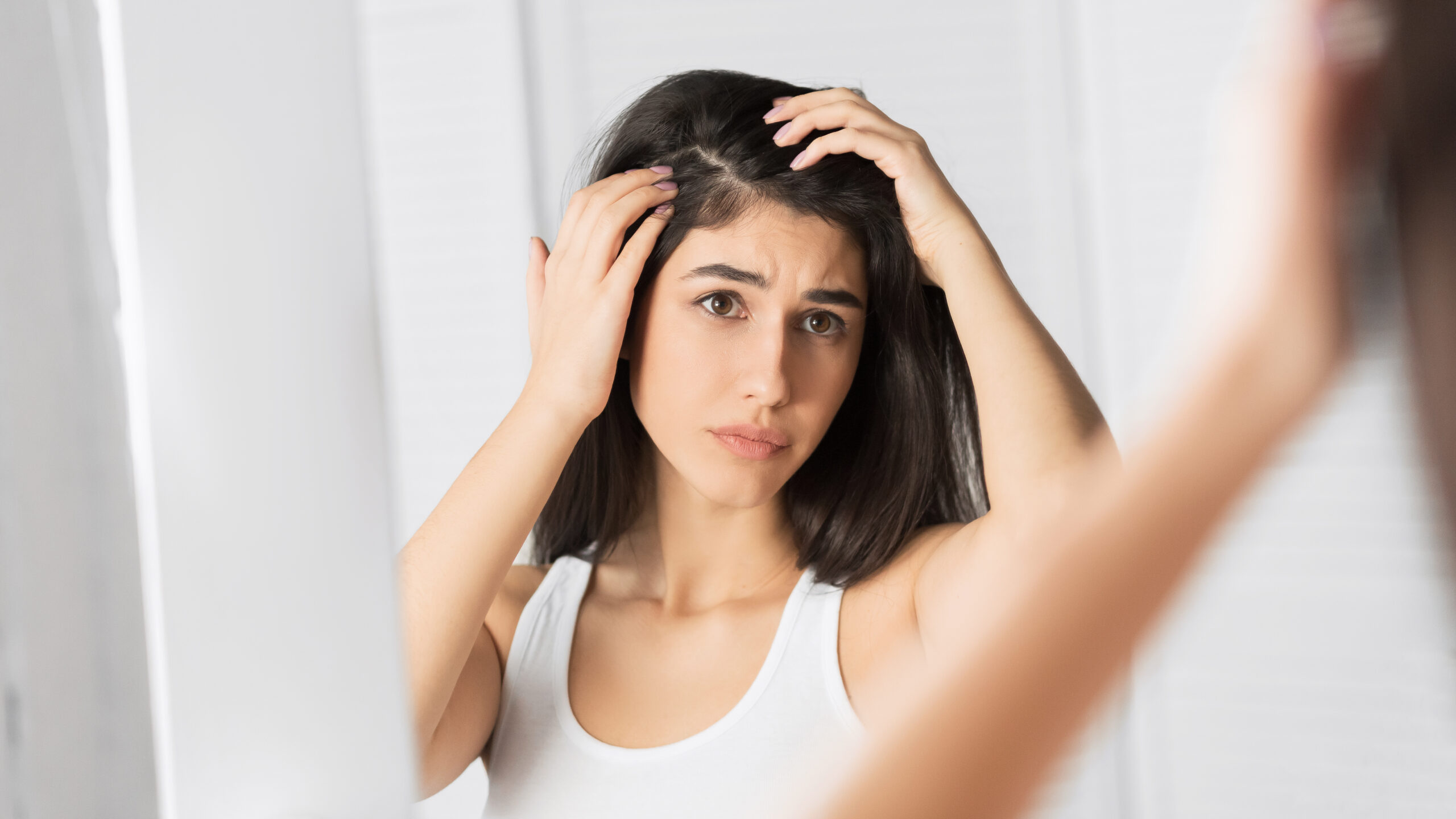 The image captures a young woman examining her scalp in front of a mirror. She uses both hands to part her hair, focusing on her hairline.