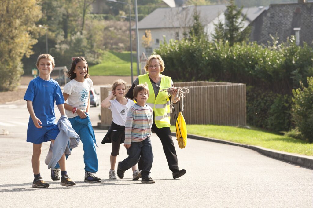 Photos d'enfants en train de marcher pour aller à l'école avec Pédibus, une dame est derrière les enfants en gilet jaune