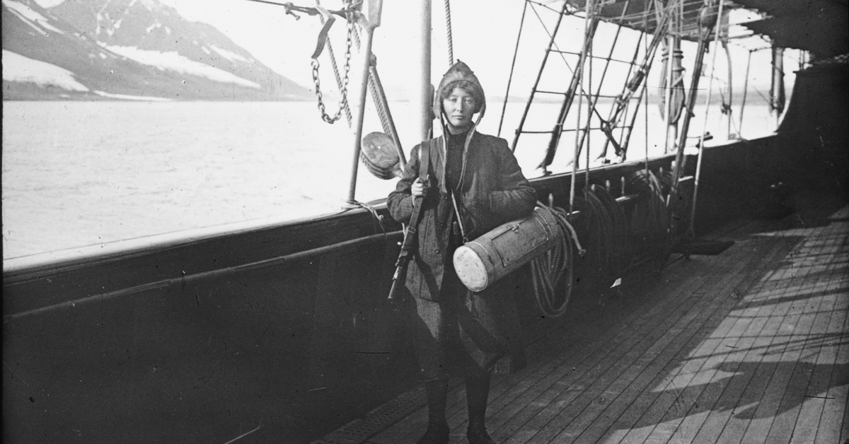 En godt kledd kvinne utstyrt med rifle og botanikerveske, ombord på en yacht ved Svalbard. Bildet er sort og hvitt, kvinnen ser bestemt ut i midten.