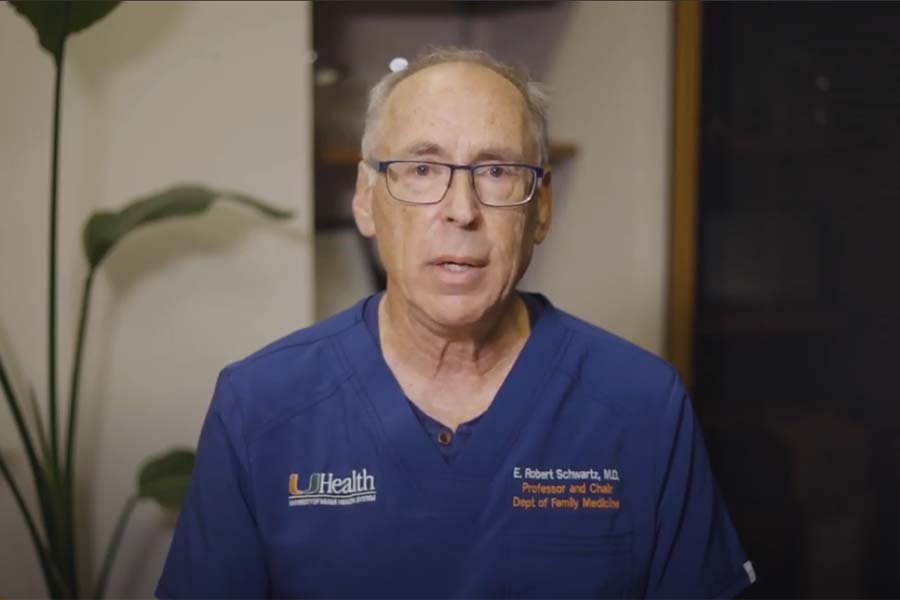 E. Robert Schwartz wearing blue scrubs and glasses, he's speaking on camera