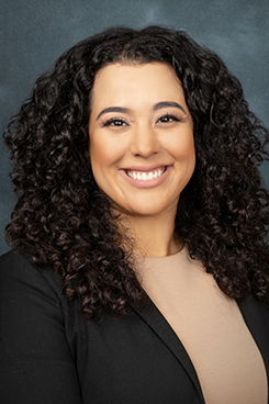 Headshot of a woman, she has long curly hair, is wearing a coat, and a brownish pink blouse