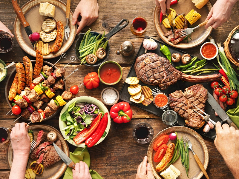 Table full of dishes with protein, meat, sausages, chicken, and some vegetables like paprika, corn, tomatoes and sauces