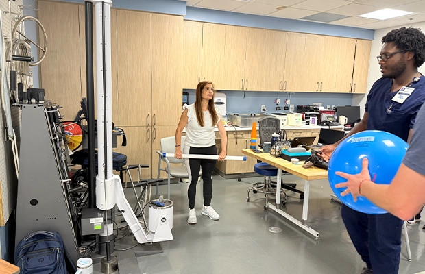 In a medical center, a woman lifts a plastic cylinder with both hands in front of two health specialists.