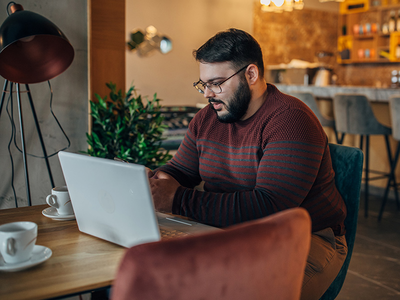 Man sitting in a cafe as he uses his phone and laptop