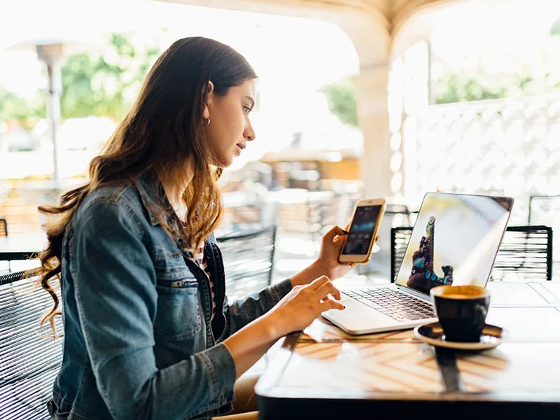 A woman sitting outdoors, she is sitting and looking at her phone and laptop