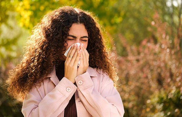 A closeup of a woman blowing her nose, she's walking outdoors surrounded by greenery