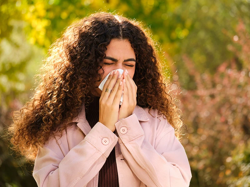 A closeup of a woman blowing her nose, she's walking outdoors surrounded by greenery